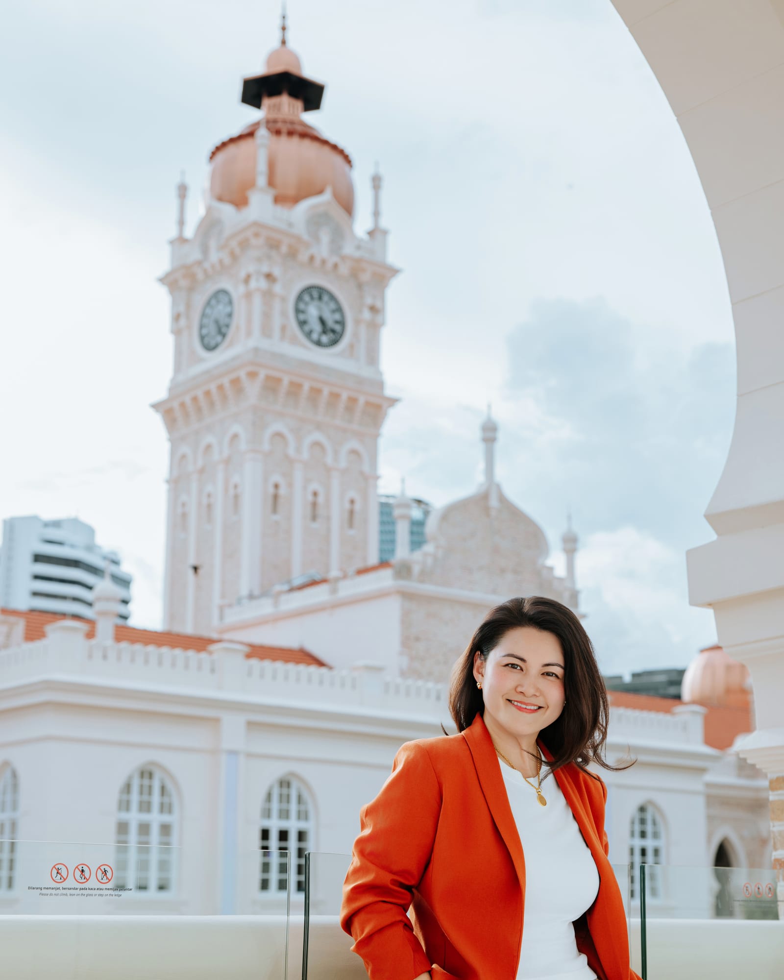 Chan Quin Er in an orange blazer, leaning on a balustrade under an arch, the clock tower of the Sultan Abdul Samad Building behind her.